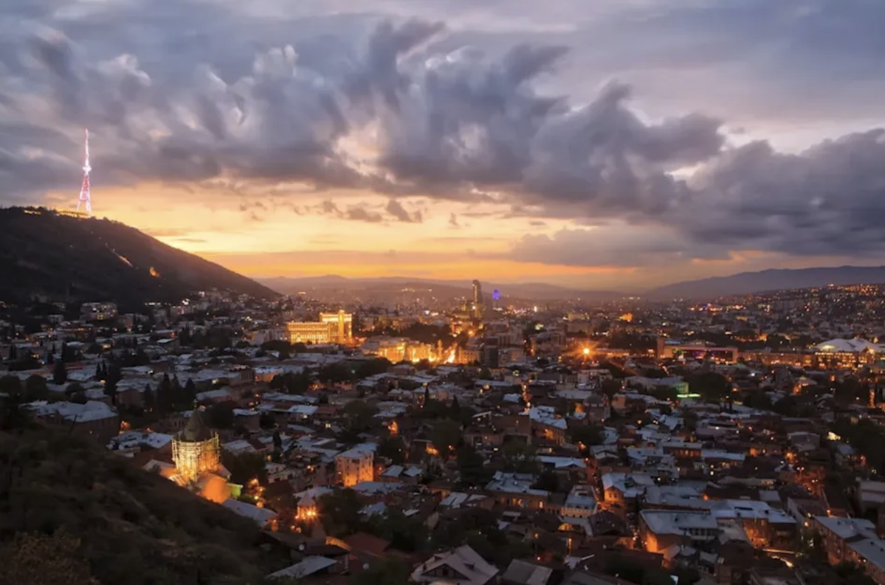 Tbilisi i solnedgång med tända stadsljus, kullar och dramatiska moln