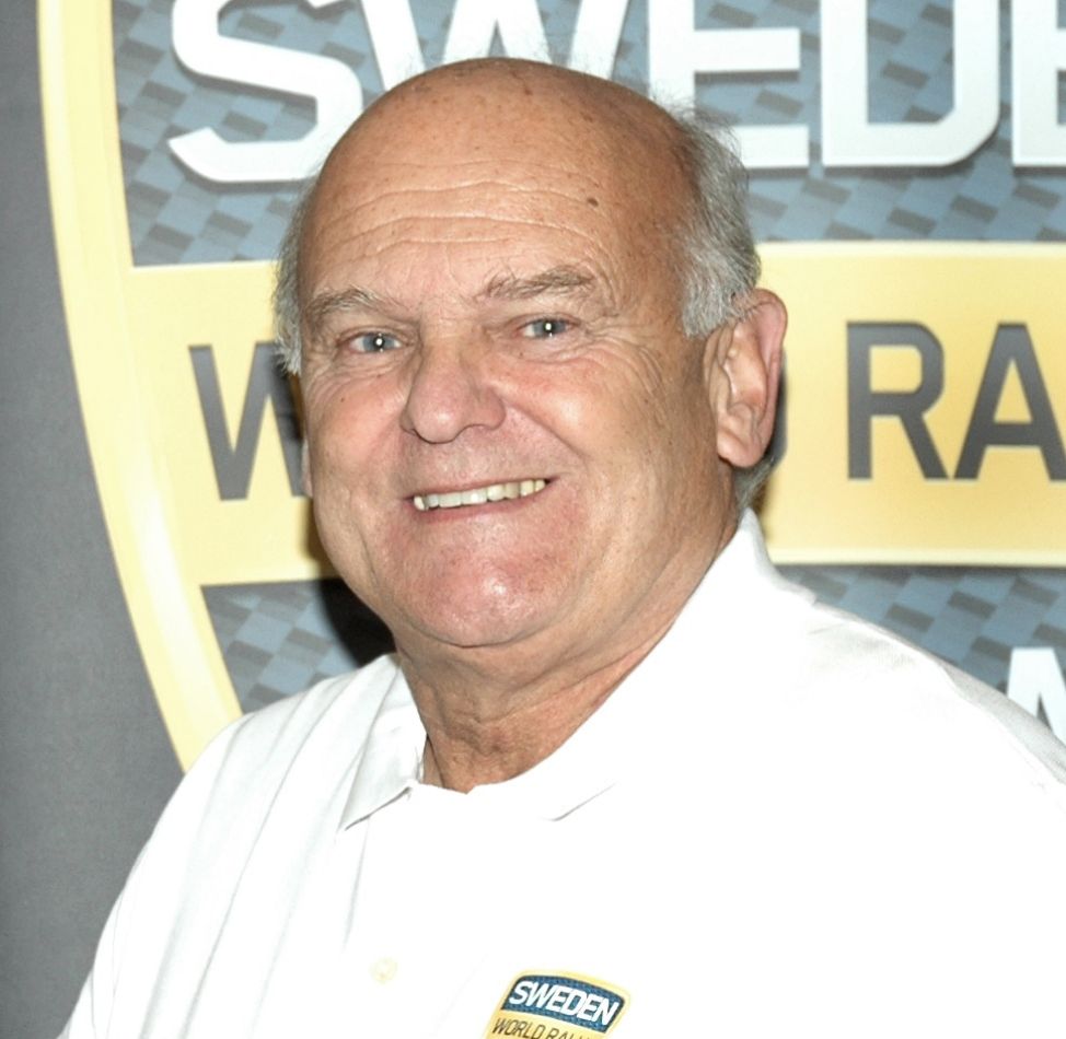 Porträtt av rallylegendaren Stig Blomqvist, leende framför en Rally Sweden-bakgrund